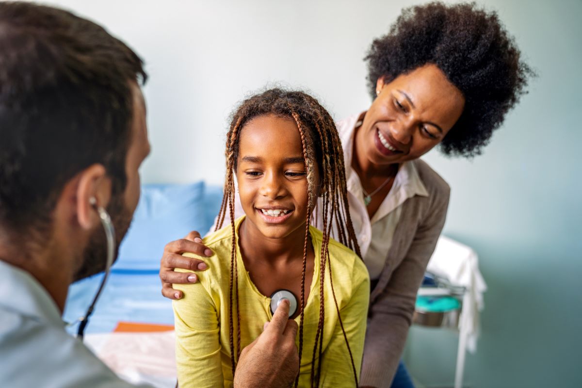 Pediatrics-Asthma-treatment Mother behind daughter holding daughters shoulder as a doctor places a listening device on a child's chest in order to hear their lungs