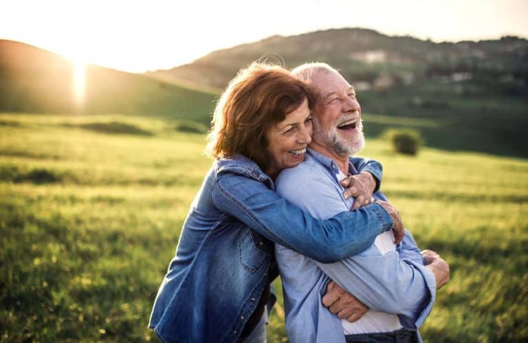 Financing The Cost of Hearing Aids Woman and man in a field with sun going down. Woman is holding man from behind in a familiar way as he laughs.