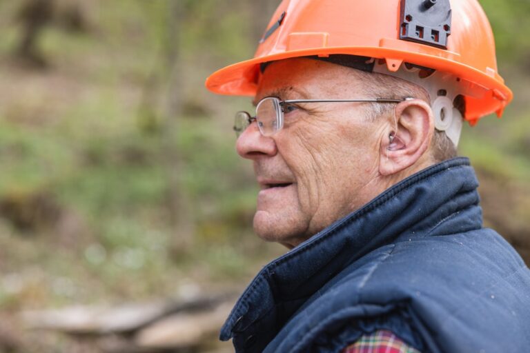 Senior male lumberjack wearing hearing aid looking away while working in forest Senior man wearing a hearing aid and hard hat