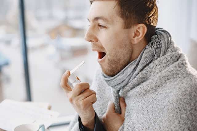 man with sore throat spray spraying does into mouth. He is wrapped in a grey blanket.