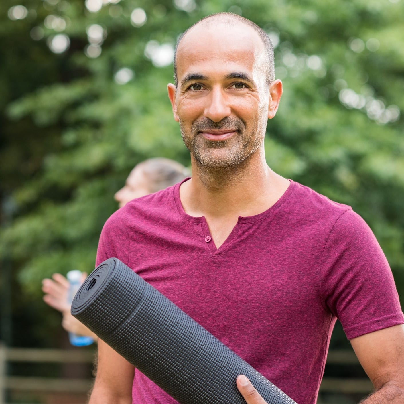 Smiling man holding a yoga mat at an outdoor yoga class.